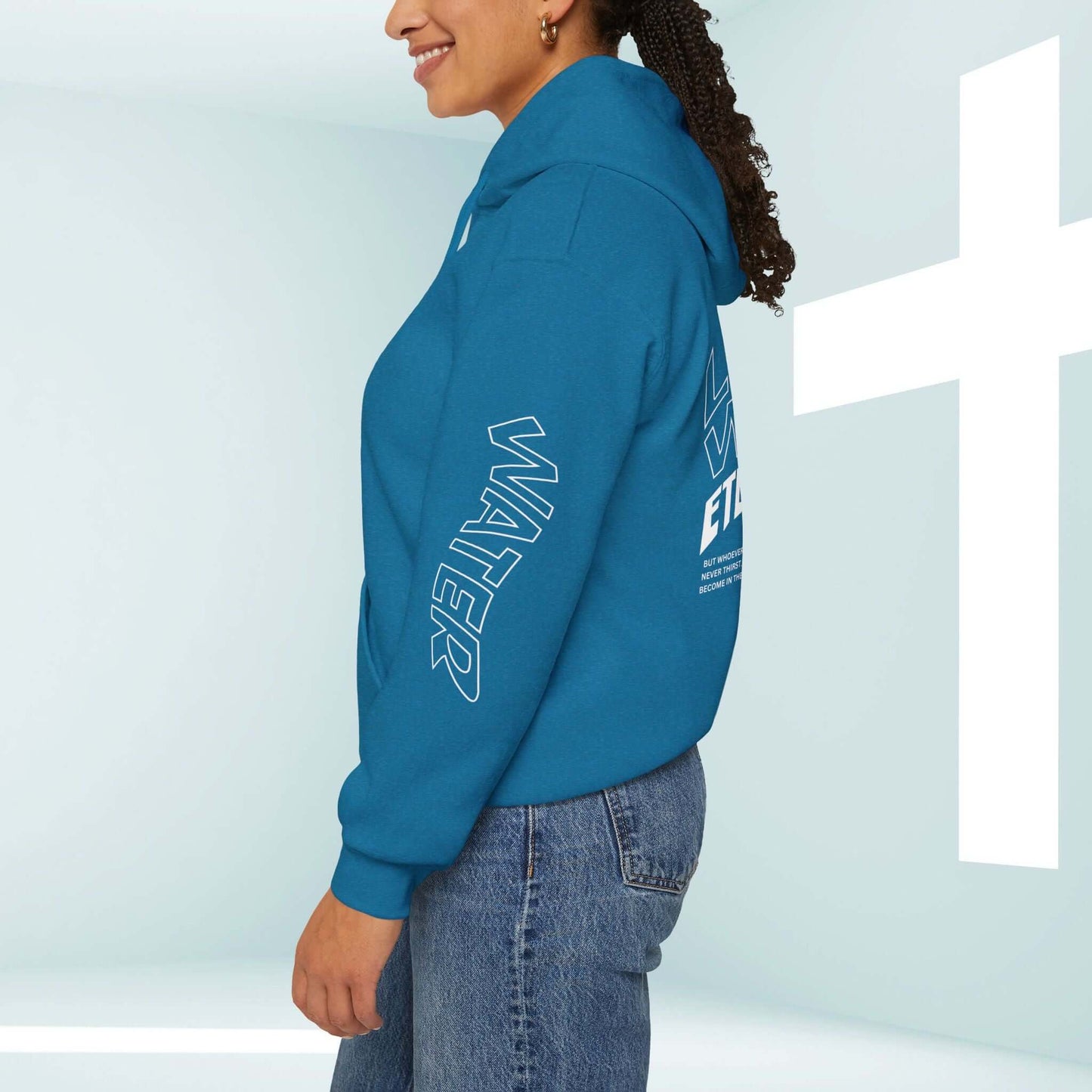 Woman wearing blue Living Water Eternal Life Christian hoodie, featuring "WATER" graphic on sleeve, standing by white cross in background.