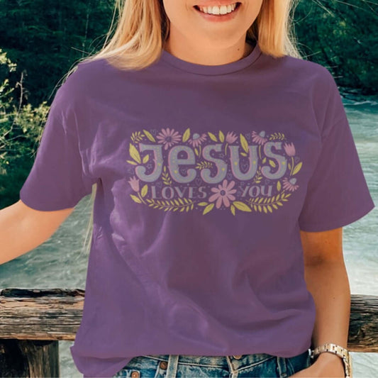 Woman wearing purple "Jesus Loves You" t-shirt amidst scenic outdoor backdrop, showcasing Christian apparel and inspirational shirt design.