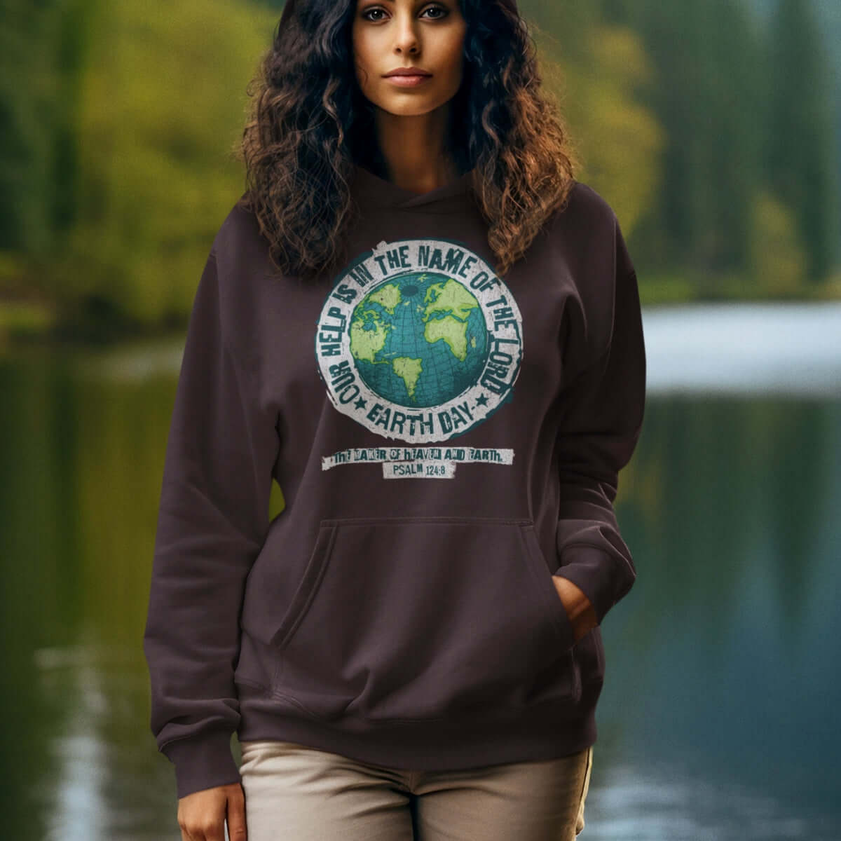 Woman wearing Earth Day "Our Help Is" Christian hoodie with Bible verse, standing outdoors near a lake, promoting faith-based and environmental message.