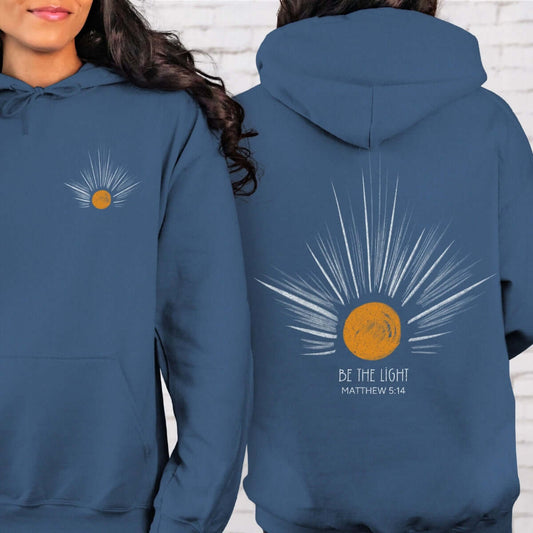 Woman wearing Be The Light Sunburst Christian hoodie with sun rays graphic, inspirational Bible verse, and celestial sun design