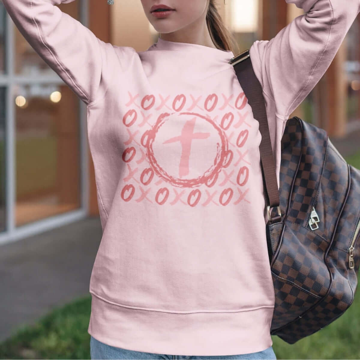 Woman wearing pink Christian wedding love sweatshirt with XOXO heart cross design Valentines Day gift, standing outside with backpack