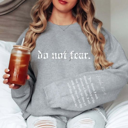 Woman wearing a gray "Do Not Fear" Christian sweatshirt holding an iced drink, sitting on a bed - no fear faith-based apparel