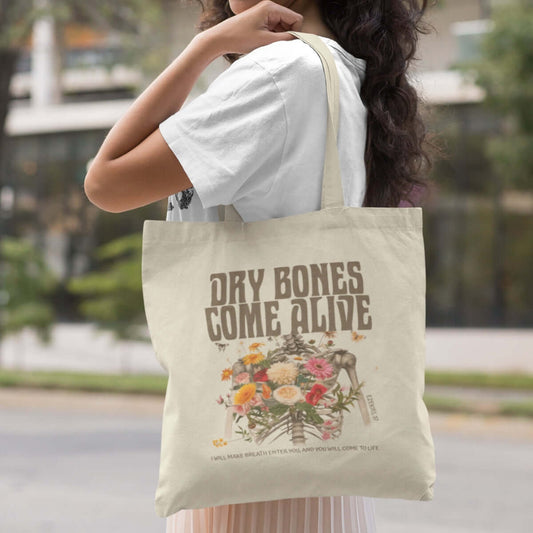 Woman carrying a Christian Halloween tote bag with "Dry Bones Come Alive" design, featuring skeleton and floral artwork.