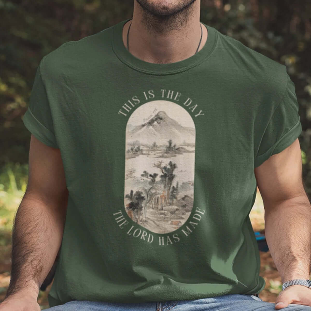 Man wearing "This Is The Day The Lord Has Made" green mountain nature camping Christian T-shirt outdoors.