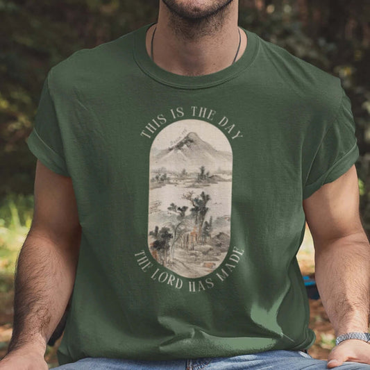 Man wearing "This Is The Day The Lord Has Made" green mountain nature camping Christian T-shirt outdoors.