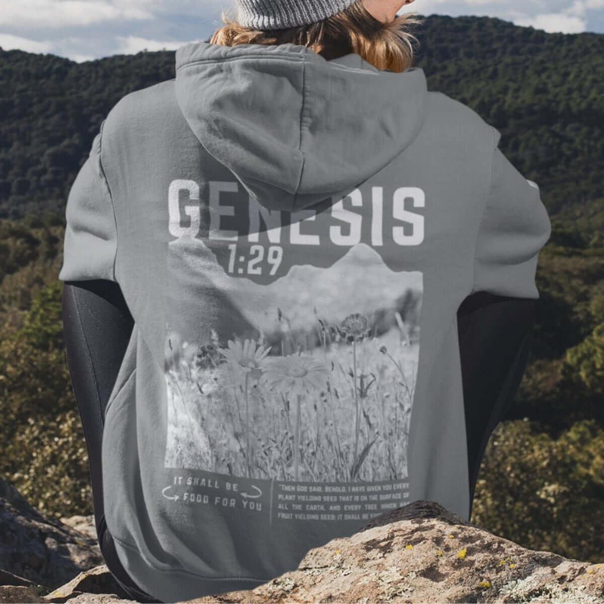 Person wearing Genesis hoodie with Bible verse, sitting on rocks overlooking nature; outdoor religious faith-based clothing.