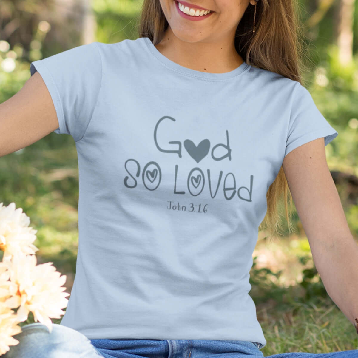 Woman wearing light blue "God So Loved" John 3:16 Christian t-shirt, sitting outdoors with flowers, spreading faith and inspiration.