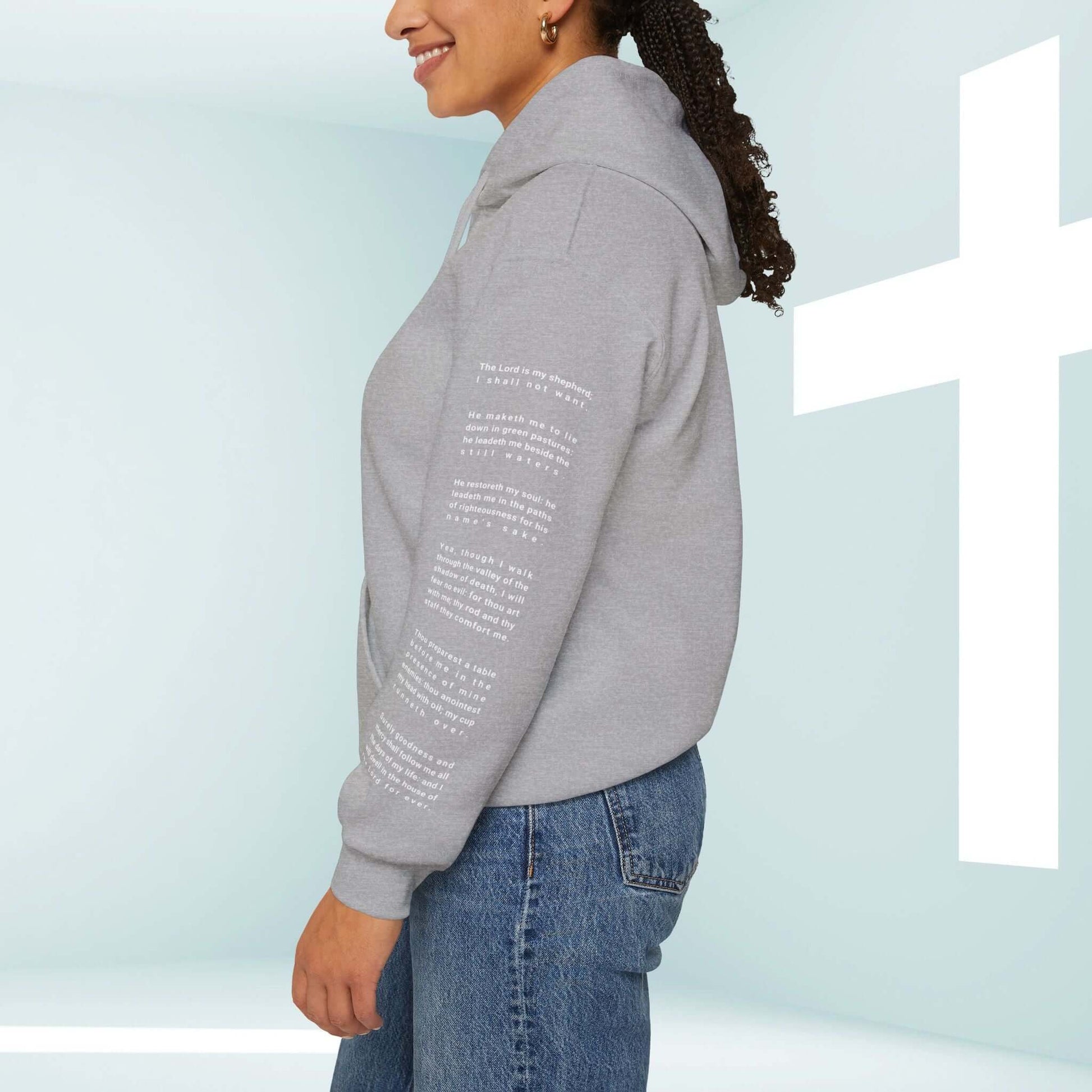 Woman wearing a grey Fearless Psalm 23 No Fear Christian hoodie with Bible verses on the sleeve in front of a cross background.