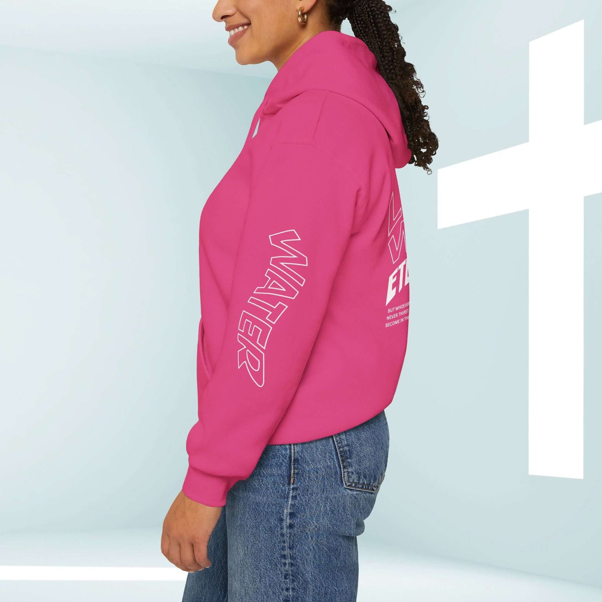 Woman wearing pink Living Water hoodie, featuring Christian graphics on sleeves and back, standing by a white cross in a bright room.