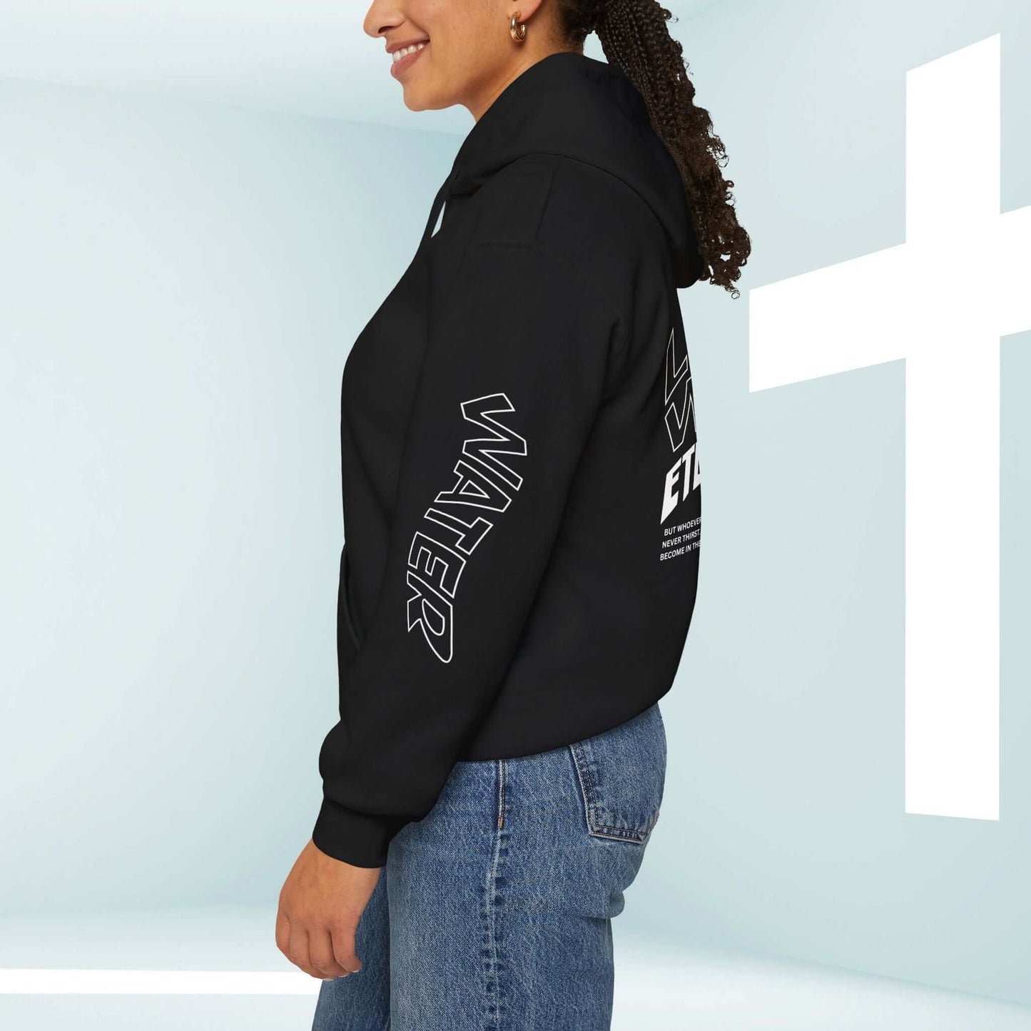 Woman wearing black Living Water Eternal Life Christian hoodie with graphics on sleeves, front, and back in front of a white cross.