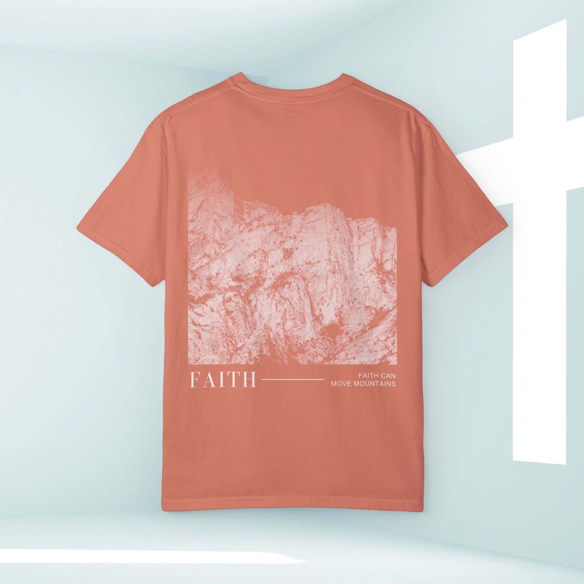 Faith Can Move Mountains Christian T-shirt with mountain graphic and faith message in coral color on display with cross in background