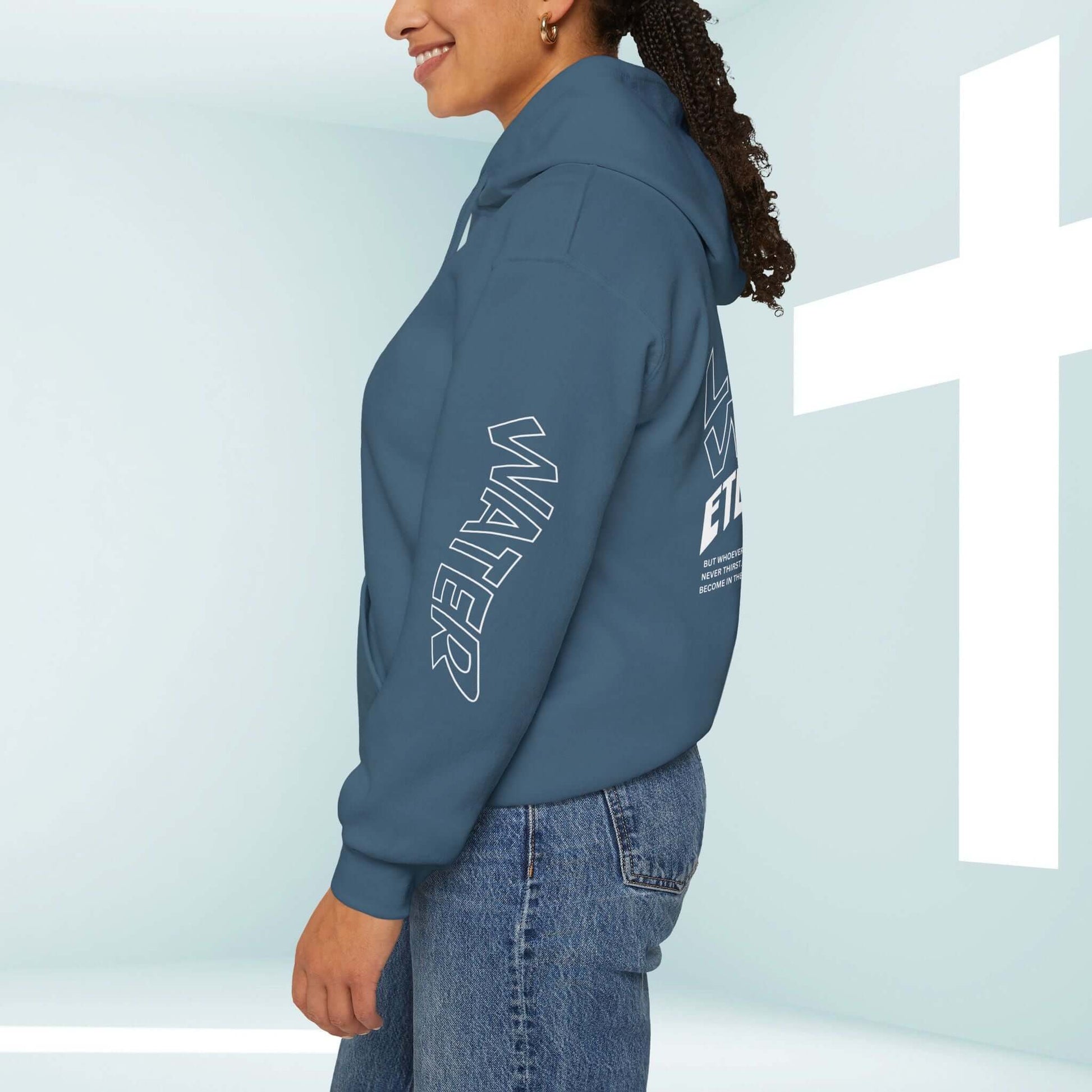 Woman wearing a blue Living Water Eternal Life Christian hoodie with sleeve and back graphics, standing next to a white cross.