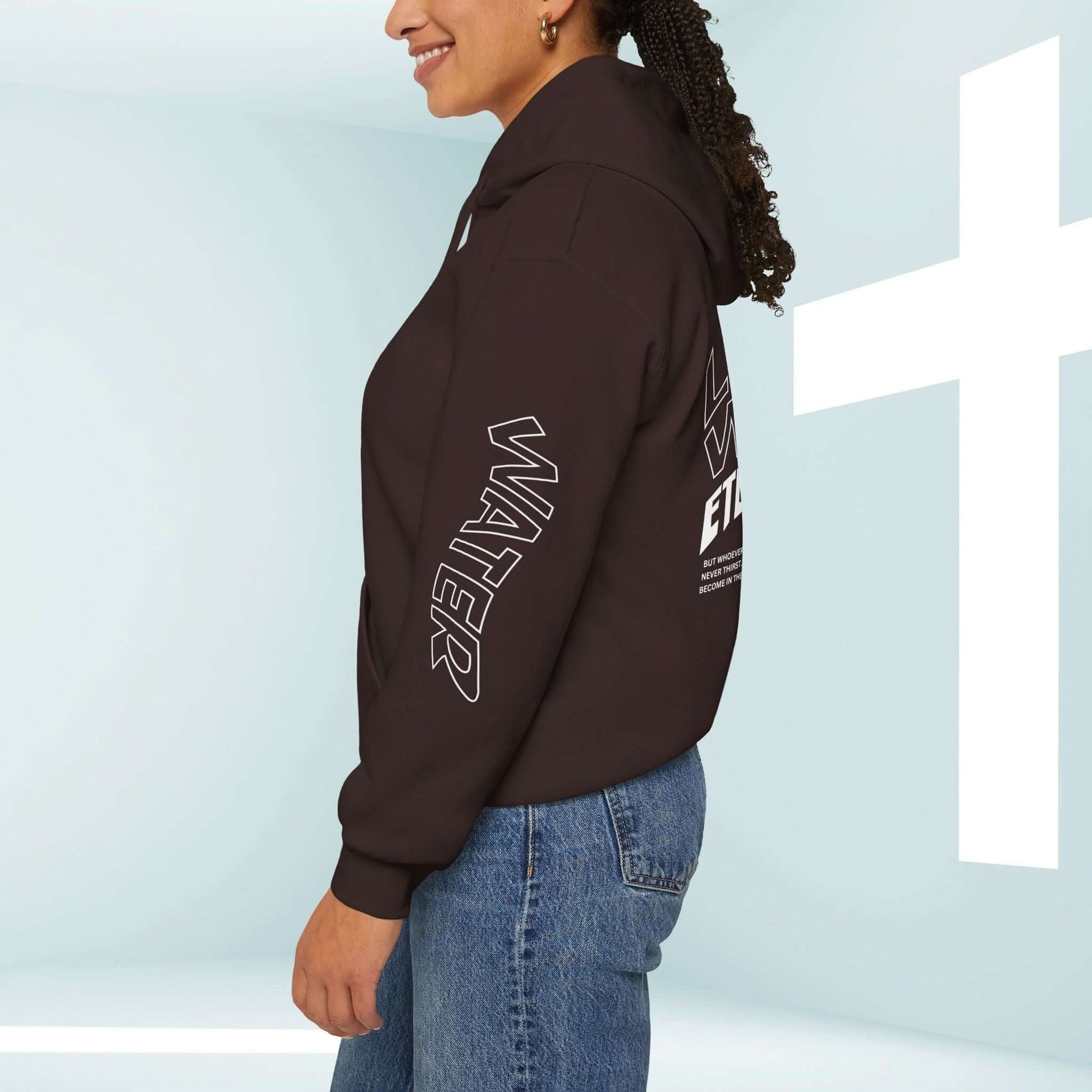 Woman wearing a black Living Water Eternal Life Christian hoodie with sleeve and back graphics, standing next to a white cross in the background.