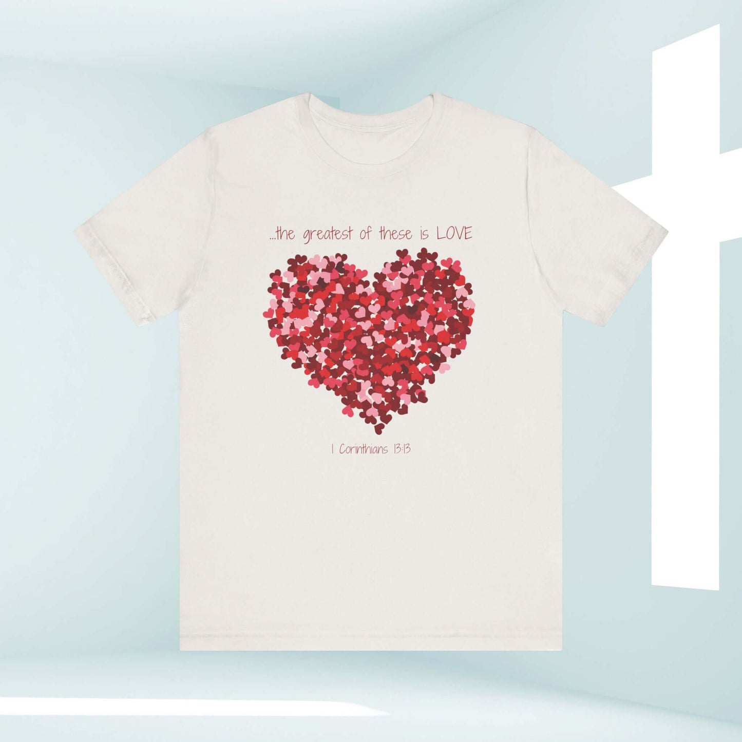 Christian Valentines Day t-shirt with heart graphic and "The Greatest of These Is Love" message in front of a white cross background.