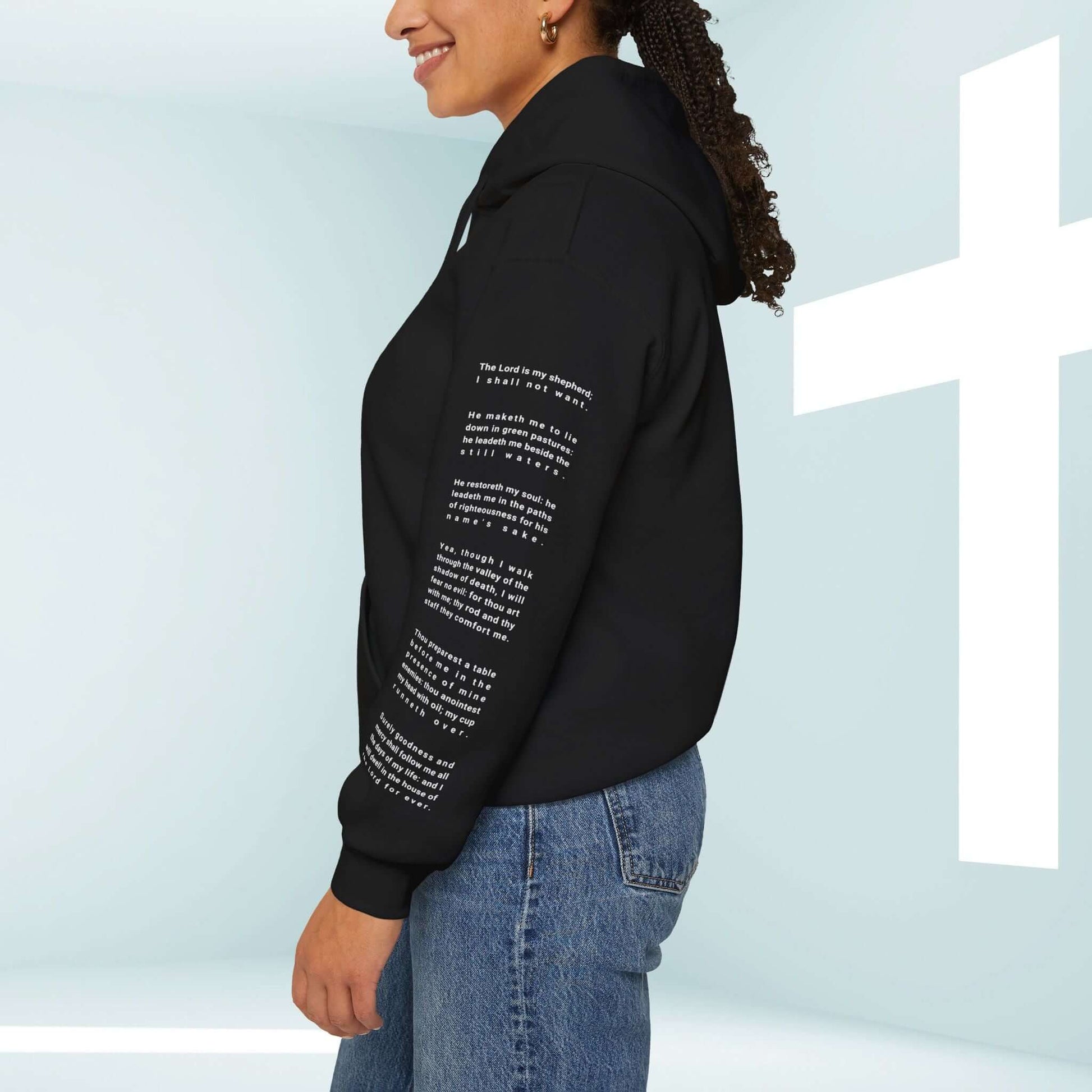 Christian woman wearing a black Fearless Psalm 23 hoodie with inspirational Bible verse graphic on the left sleeve, standing near a white cross.