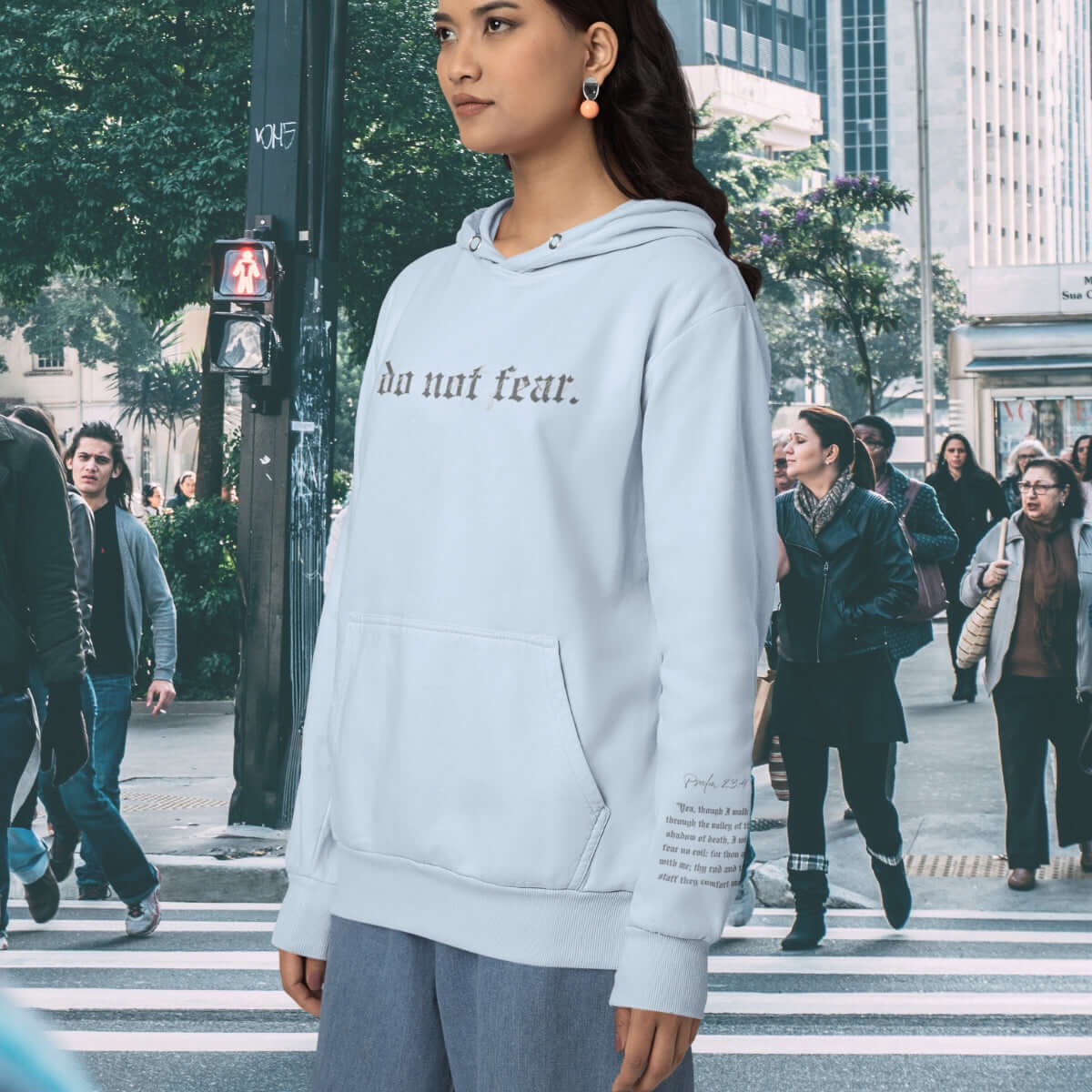 Woman wearing light blue Christian hoodie with Bible verse, standing in front of a cross, promoting faith-based apparel and inspirational clothing.