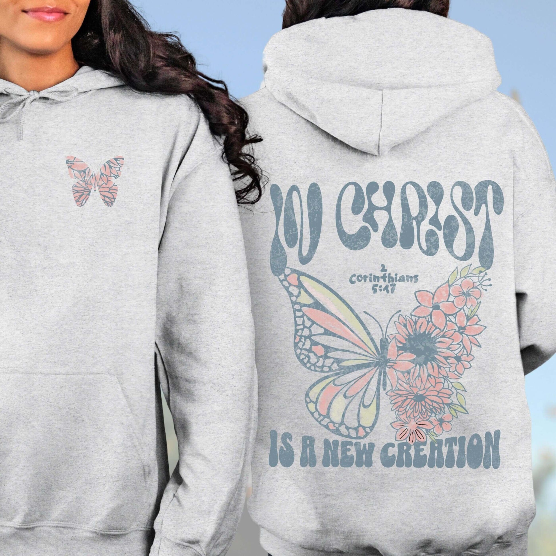 Woman wearing gray Christian hoodie featuring "In Christ Is A New Creation" with butterfly and flowers, inspirational faith-based sweatshirt