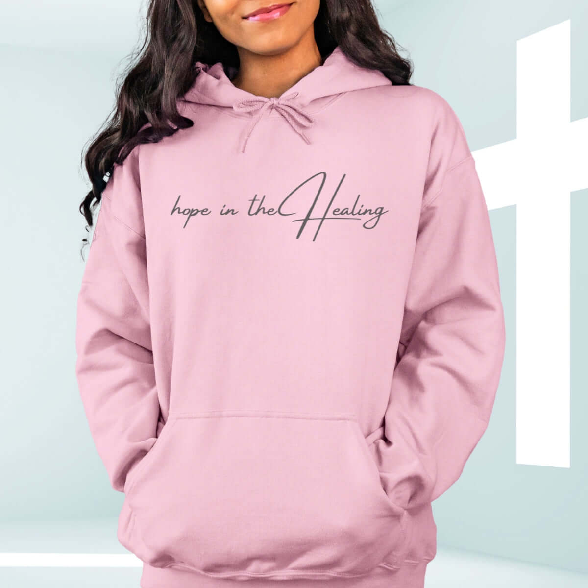 Light pink "Hope In The Healing" Christian hoodie on white wooden background with flowers.