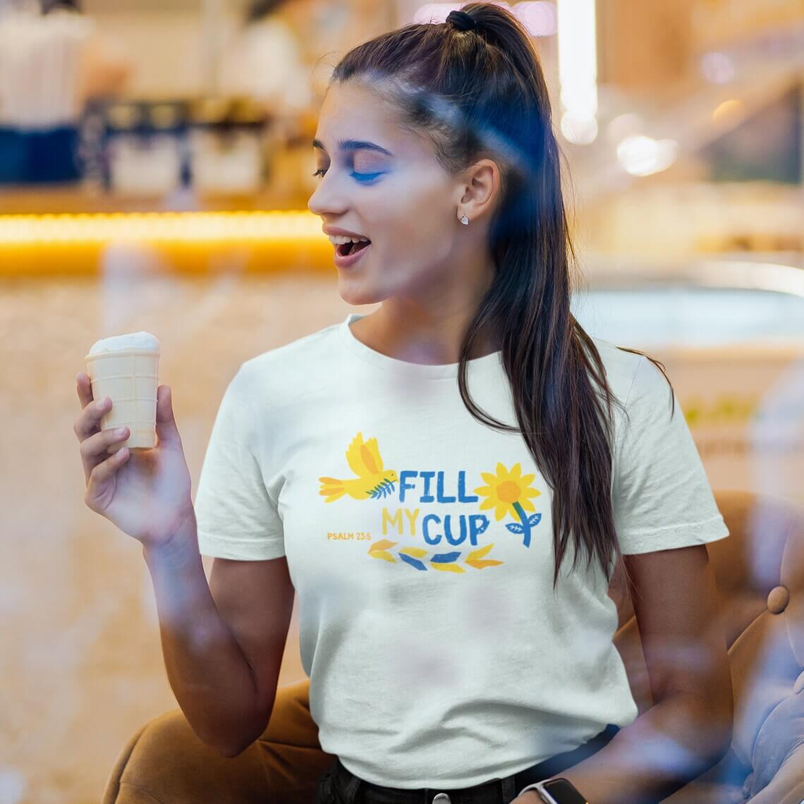 Woman wearing Floral Fill My Cup Bird and Flower Christian T-Shirt while enjoying a drink in a cozy café setting.