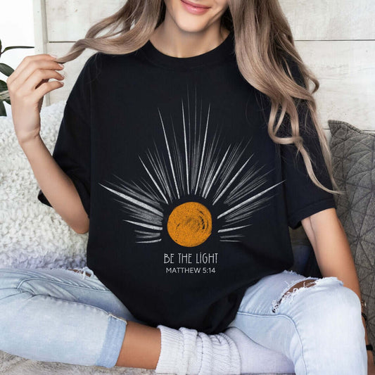 Woman wearing Be The Light sunburst Christian t-shirt with Matthew 5:14 Bible verse, celestial sun ray design, faith-inspired clothing.