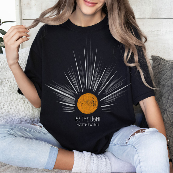 a person wearing a black Christian t-shirt with an image of the sun on it saying "Be The Light"